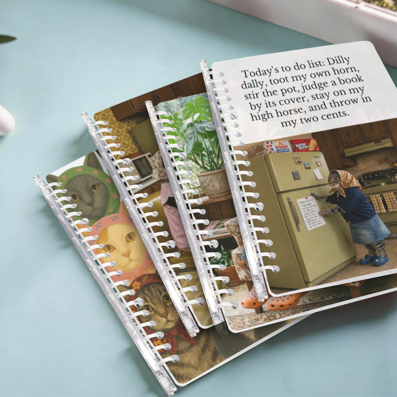 Three spiral-bound mini notebooks with printed cartoon and lifestyle illustrations are arranged on a light blue surface. The top notebook is open, showing a white page with a black handwritten-style list that reads, 'Today's to do list: Dilly dally, toot my own horn, stir the pot, judge a book by its cover, stay on my high horse, and throw in my two cents.